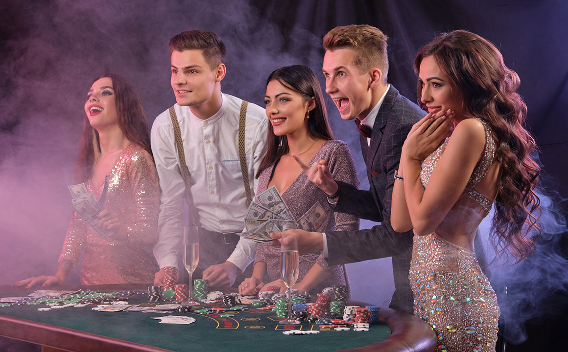 Friends playing poker at casino, at table with stacks of chips, money, cards, champagne on it. Celebrating win, excited. Black background. Close-up.